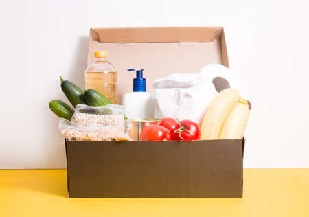 box with products for donation, yellow background, copy space, cereals, vegetables, fruits, oil in a bottle, flour and canned food in a box, hygiene means of protection against coronavirusの写真素材