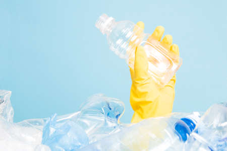 a hand in a rubber yellow glove holds an empty plastic bottle on a blue background, a hand in a pile...