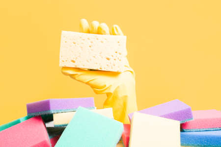 a hand in a yellow rubber glove holds a sponge for washing dishes and cleaning, a hand sticks out of a pile of sponges, yellow background copy spaceの写真素材
