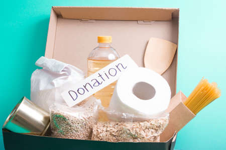 donation inscription on a white cardboard box, food box on a blue-green background, copy space, toilet paper, spaghetti, vegetable oil, cereals, canned foodの写真素材
