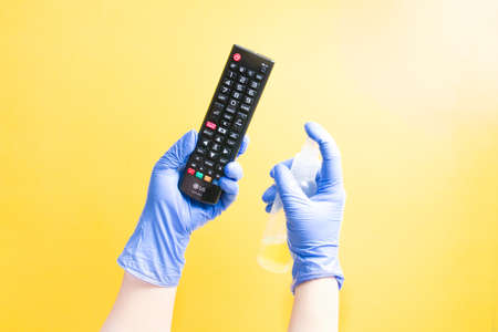 a hand in a one-time rubber glove is spraying a TV LG remote control with an alcohol sanitizer, yellow background, copy spaceの写真素材