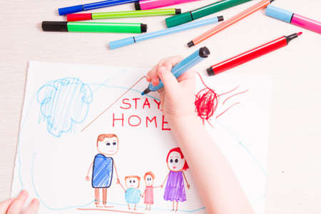 the child draws with felt-tip pens a family and a house on paper, felt-tip pens on the table, the inscription in the picture stay home, quarantine concept, self-isolation with the childの写真素材