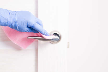 a hand in a blue disposable rubber glove wipes a doorknob with a pink rag, cleaning and disinfecting surfaces, the safety of your home, copy space, metal handle, light wooden doorの写真素材