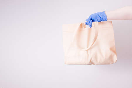 a hand in a blue disposable medical rubber glove holds a shopping bag for shopping, light background, copy space, contactless delivery concept, self-isolation and quarantine, light backgroundの写真素材