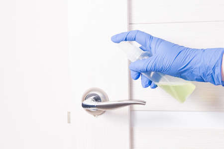 a hand in a blue disposable rubber glove sprinkles with an alcohol disinfectant a door handle, a...