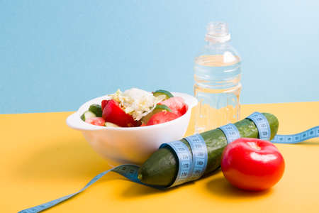 small bottle of water, salad in a white bowl, blue measuring tape around a cucumber, tomato on a...