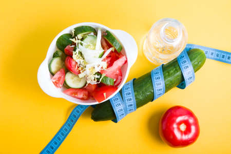 small bottle of water, salad in a white bowl, blue measuring tape around a cucumber, tomato on a blue and yellow background copy space, diet and weight loss concept, top viewの写真素材