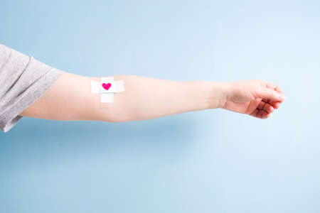 female hand with a band-aid glued with a cross on a vein, heart on a band-aid, blood donor concept, blue background, copy spaceの写真素材