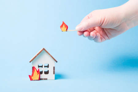 burning wooden model of a house, fire cut out of felt, blue background copy space, insurance and...