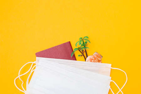 toy palm, red passport and a shell under a medical protective disposable face mask, yellow background copy space, travel and quarantine cancellation of vacation and beach holidaysの写真素材