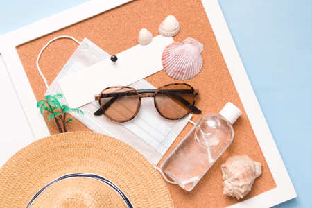 straw hat, gel sanitizer, medical face mask, palm tree, sunglasses and cockleshells on cork board, blue background, copy space, top view, travel and quarantine conceptの写真素材