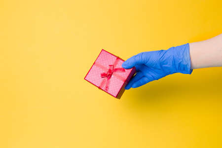 a hand in a blue disposable glove holds a red gift box with a bow from a red ribbon with gold thread, yellow background, copy spaceの写真素材