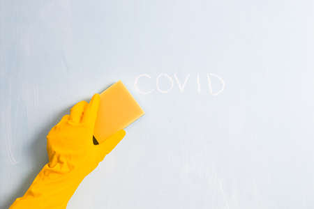 hand in a rubber yellow glove wipes the word covid with a sponge, light blue background, copy space,...