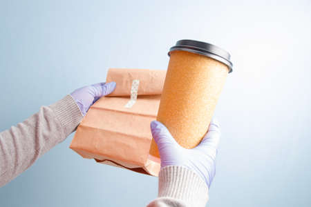 female hands in blue rubber disposable gloves hold a large paper cup of coffee and a paper bag, blue...