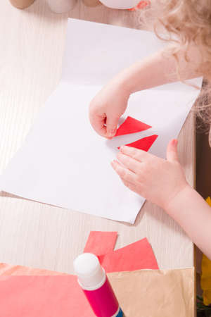 a small child glues paper, children's safe scissors and colored paper on the table, what to do with the child at home, children's hands glue colored triangles on a sheet of white paperの写真素材