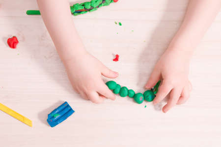 a small child sculpts a caterpillar from green plasticine on a wooden table, the development of fine motor skills of hands, what to do with the child at home, stay at homeの写真素材