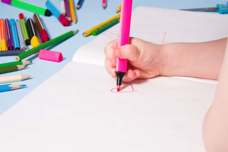 a small child draws with a pink felt-tip pen a heart in an album, copy space, top view, blue...