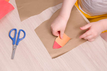 small child glue a house of paper, children's safe scissors on a wooden table, what to do with the child at home, copy space, children's hands on the tableの写真素材