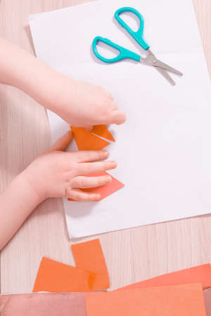 a small child glues paper, children's safe scissors and colored paper on the table, what to do with the child at home, children's hands glue colored triangles on a sheet of white paperの写真素材