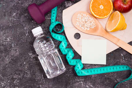 bottle of water, dumbbells, measuring tape, diet food on a black background, sticker for your text, vegetables, fruits, oatmeal on a wooden spoon, healthy lifestyle conceptの写真素材