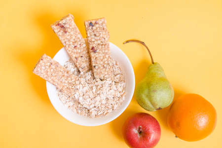 several muesli and fruit bars in a bowl with dry oatmeal and various fruits on a yellow background,...