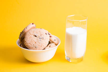 glass of milk and cookies with pieces of chocolate and lingonberries in a white bowl on a yellow...