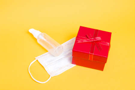white protective medical children's mask and gel antiseptic next to a red gift box with a bow on a yellow background copy space, holiday in quarantine conceptの写真素材