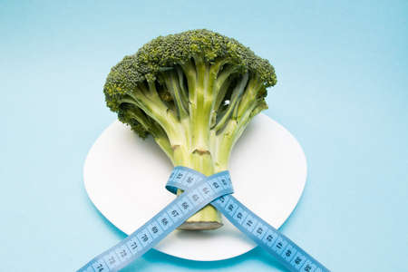 head of broccoli on a white plate wrapped in blue measuring tape, slimming and weight loss concept /...