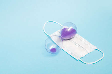 broken hourglass with purple sand and a medical protective white mask on a blue background copy...