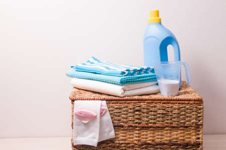 clothes in light blue tones, strange powder in a measuring cup and gel for washing colored clothes in a blue plastic bottle on a wicker laundry basket, children's socks stick out of the basketの写真素材