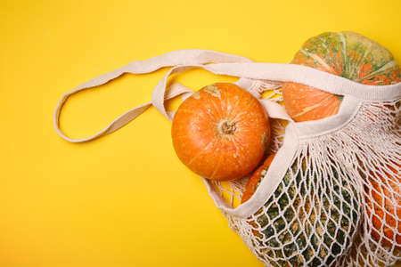 Fresh pumpkins, pattypan squash in shopping eco-friendly mesh bag, autumn leaves on yellow...