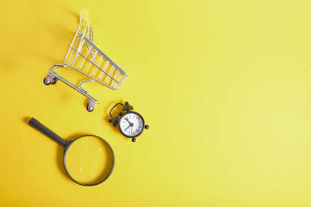 magnifier, alarm clock and miniature shopping basket on yellow background top view copy spaceの写真素材