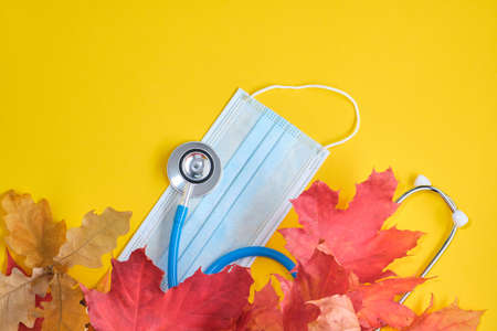 disposable protective medical mask and stethoscope and autumn maple leaves on yellow background copy spaceの写真素材