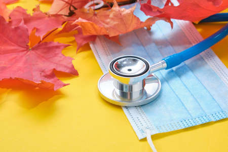 disposable protective medical mask and stethoscope and autumn maple leaves on yellow backgroundの写真素材
