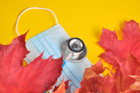 disposable protective medical mask and stethoscope and autumn maple leaves on yellow backgroundの写真素材