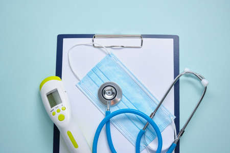 medical face mask, non-contact thermometer, paper holder with empty white sheets, medical stethoscope, pills on a blue background ,, copy spaceの写真素材