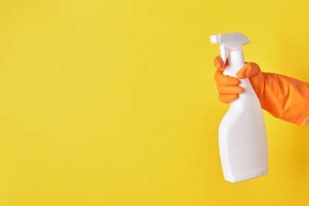 A hand in a orange glove holds a white spray bottle of cleaning fluid on a yellow background