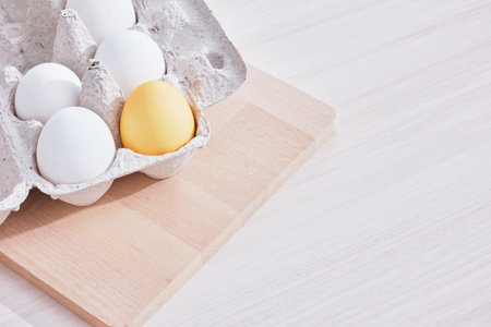 Minimal concept Easter. one colored egg and several white eggs in a cardboard box and a wooden cutting board on the tableの写真素材