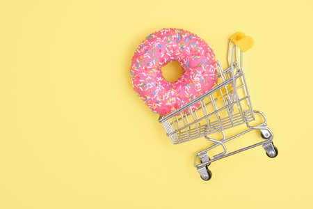 Donut with sprinkles and pink icing in a shopping cart on yellow background top view copy space