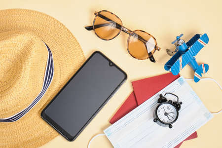 face mask, straw hat, smartphone, sunglasses, alarm clock and passport on beige background, travel during lockdown, safe beach vacation conceptの写真素材
