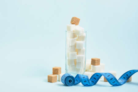 measuring tape and cubes of white and brown sugar in a glass and on a light blue and beige background copy space, sugar in drinks and food conceptの写真素材
