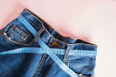blue measuring tape is threaded into jeans instead of a belt, blue jeans on a pink background, copy space, weight loss and weight control conceptの写真素材