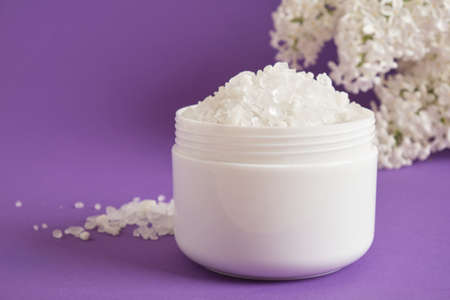 white sea salt in plastic jar and white lilac flowers on lilac background natural body care cosmetic conceptの写真素材