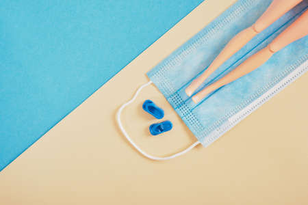 toy beach slippers and doll feet on a protective face mask on a blue and beige background, beach vacation and tourism during the epidemic conceptの写真素材