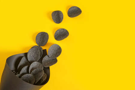 black potato chips in a black paper package on a yellow background, copy space, top view, chips made...