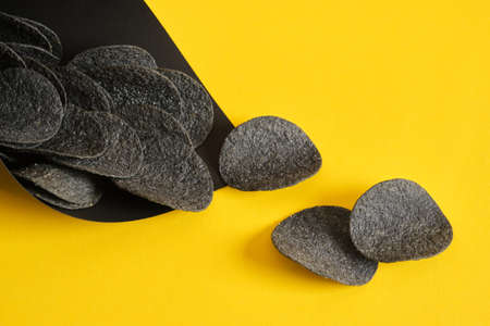 black potato chips in a black paper package on a yellow background, copy space, top view, chips made...