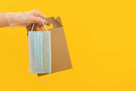 a hand in a disposable rubber glove holds a cardboard box and a face mask on a yellow background,...