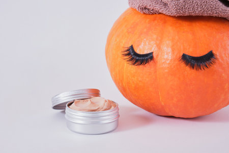 aluminum jar with cosmetic clay face mask and orange pumpkin with towel and false eyelashes on gray background, natural skin care in autumn conceptの写真素材