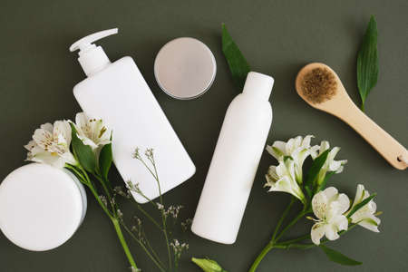 flowers, massage wooden brush, different cosmetic jars and three different blank white bottles on a dark green background backdrop Natural cosmetic packaging. mock up beauty product concept.の写真素材