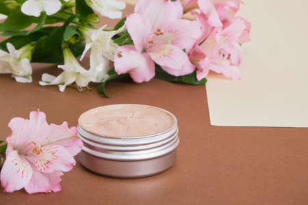 blank mock up aluminum cosmetic cream jar and beautiful pink and white lily flowers on beige brown...